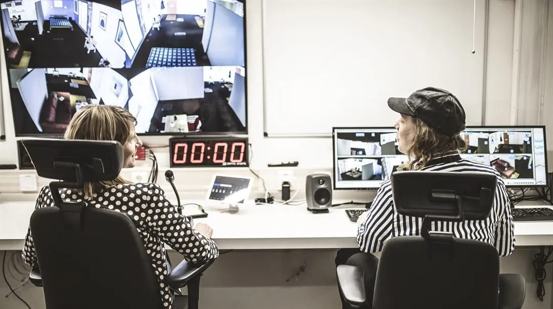 Två personer sitter vid ett långt arbetsbord. Framför dem finns flera datorskärmar, mikrofon och annan teknisk utrustning.