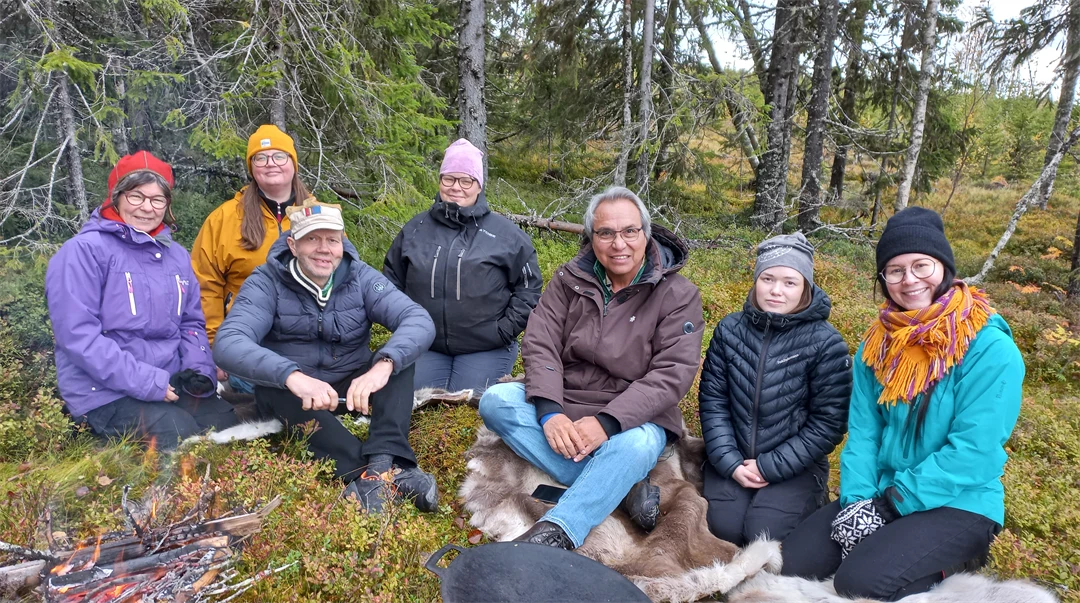 Sju personer sitter på renskinn invid en eld i skogen.