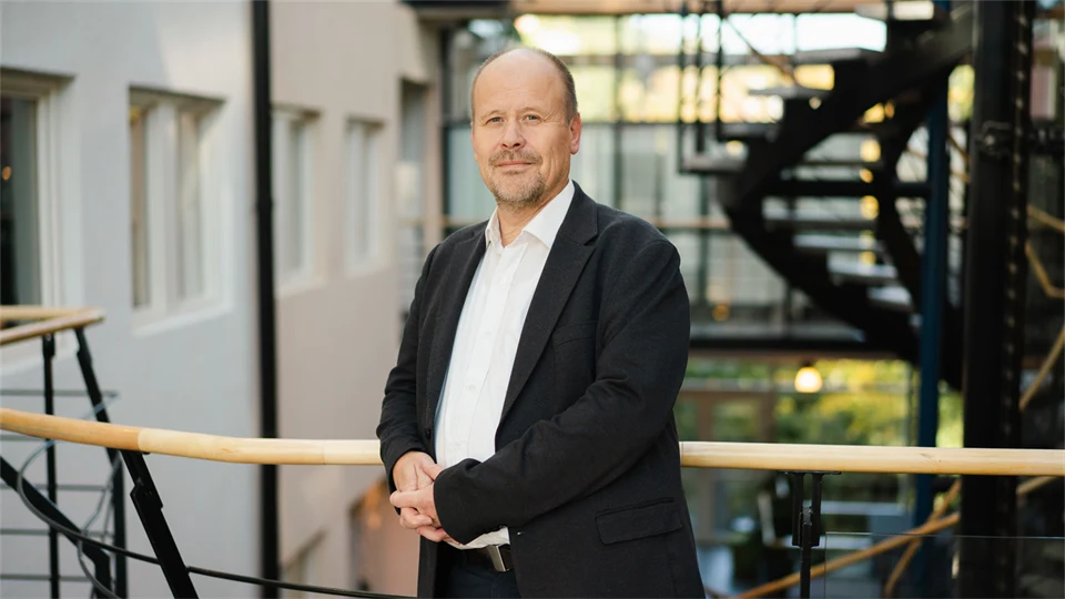 Man i vit skjorta och svart kavaj med knäppta händer tittar in i kameran. 