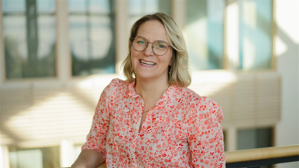 : Glad kvinna med blont hår, blommig blus och glasögon ler mot kameran. 