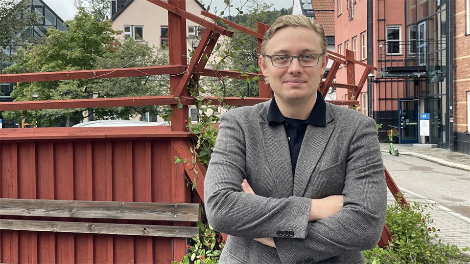 A fair-haired man with glasses and a blazer stands in front of a red fence and poses for the camera.