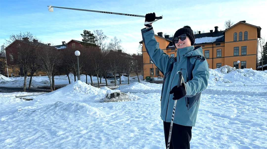 Young man with poles in his hands stands on a snow-covered field and raises one hand in the air and smiles