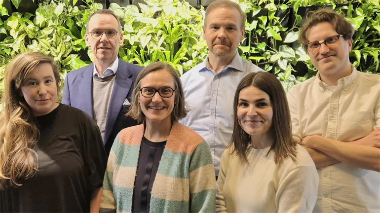 Tre män och tre kvinnor står i grupp framför gröna buskar.