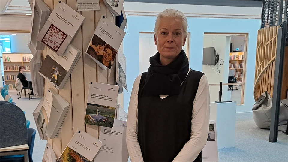 Ljushårig kvinna klädd i vit långärmad tröja med en svart väst över, runt halsen har hon en svart halsduk.