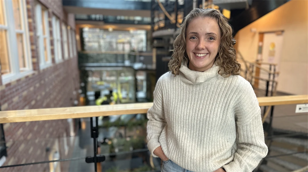 Ung kvinna med blont, lockigt, axellångt hår klädd i vit, stickad polotröja och ljusa jeans står i en ljus byggnad med många fönster, ett räcke och en trappa bakom sig. 