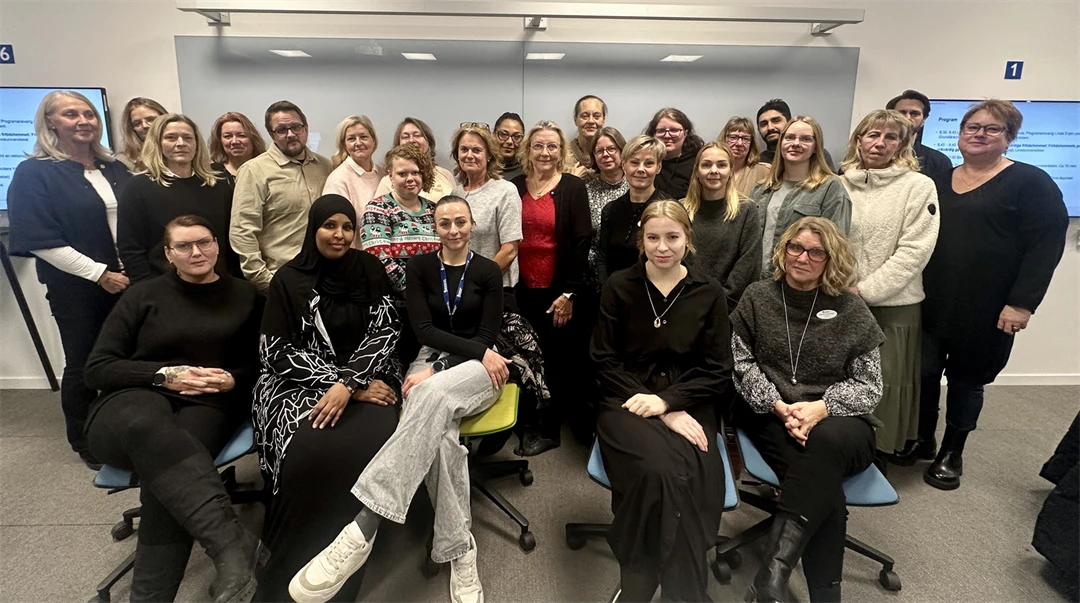 Ett trettiotal personer står i klassrum med en whiteboardtavla i bakgrunden och tv-skärmar på vardera sida. 