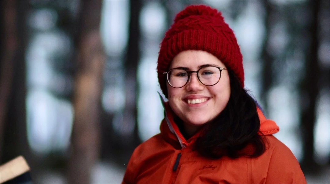 A woman with a red hat and dark brown hair wearing an orange winter jacket smiles at the camera. 