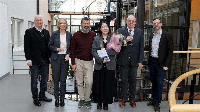 Sex personer står bredvid varandra på rad, håller glas och ser glada ut. Personen i mitten håller dessutom en blombukett och en avhandling.