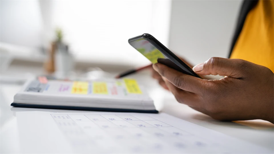 En hand håller i en mobiltelefon och en kalender ligger på ett bord.