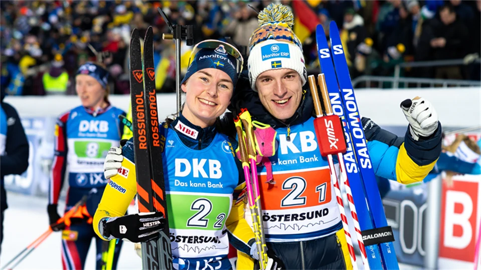 Två skidåkare bredvid varandra med tävlingskläder och skidor lutade mot axeln.