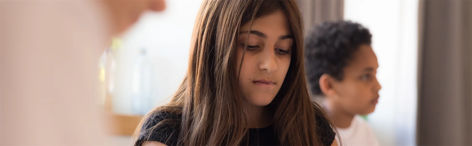 En mörkhårig flicka klädd i en svart kortärmad tröja med paljettryck sitter vid ett bord i klassrumsmiljö och håller ett papper i händerna. Snett bakom henne sitter en pojke och snett framför henne, i förgrunden, sitter ännu en pojke.  