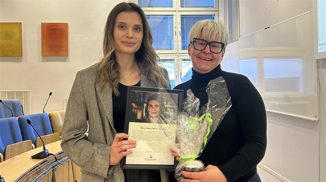 Två kvinnor står tillsammans i en föreläsningssal och håller en blombukett och en plakett i handen.