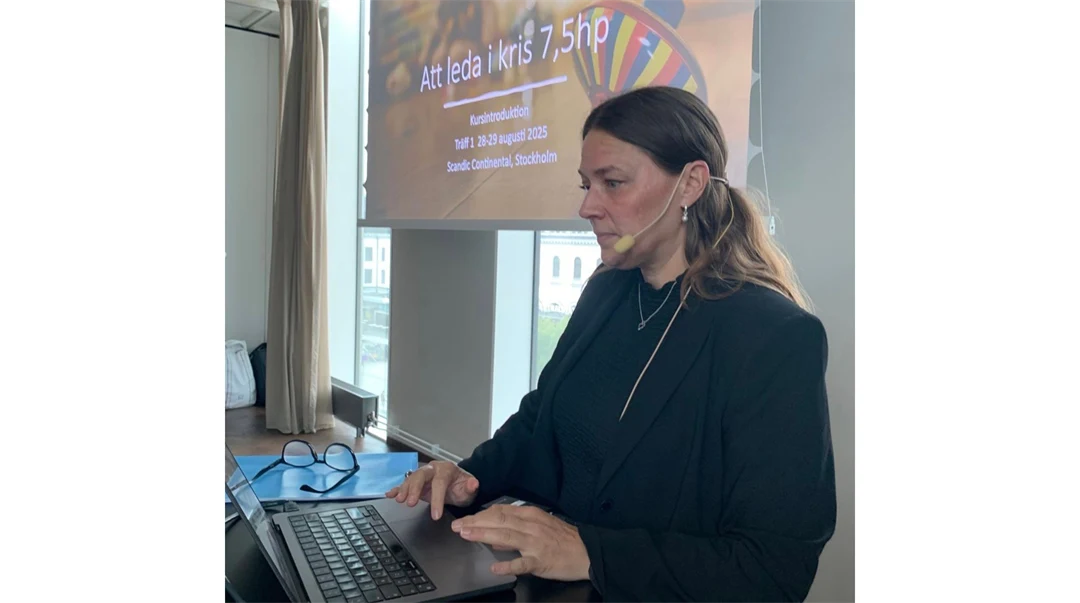 En kvinna med mikrofon står vid en bärbar dator med en skärm snett bakom sig. 