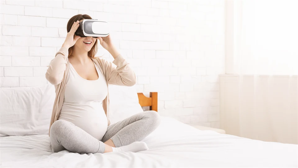 Young pregnant woman using virtual reality goggle, sitting on bed at home.