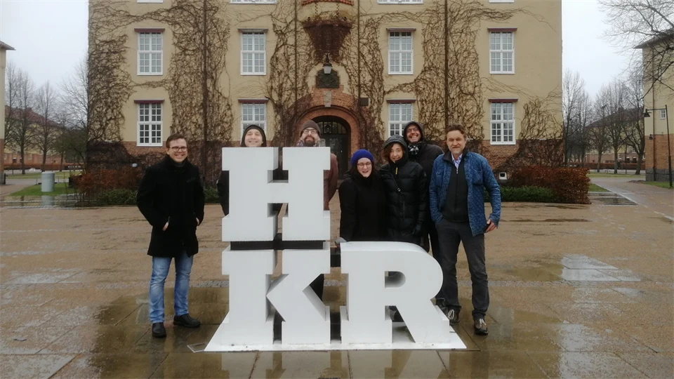 A group of people stand behind an artwork depicting the letters HKR with a large building in the background. 