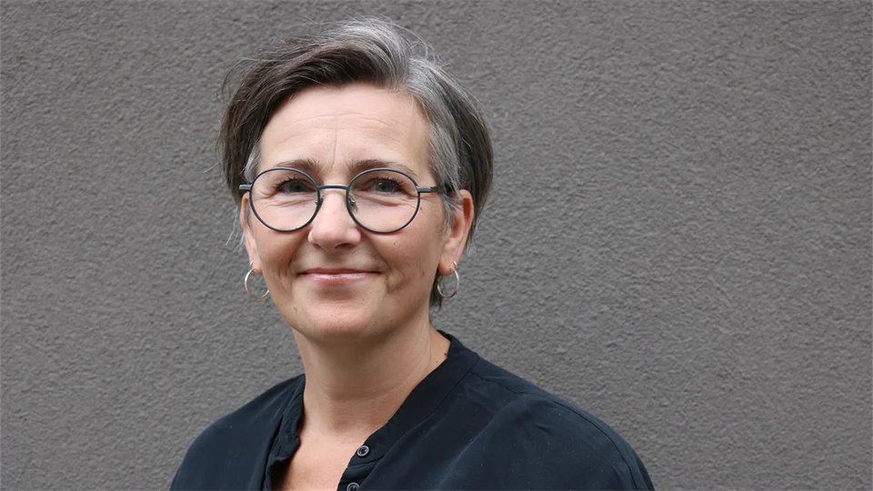 En kvinna med mörkt hår, glasögon och en svart blus står framför en mörkgrå vägg.