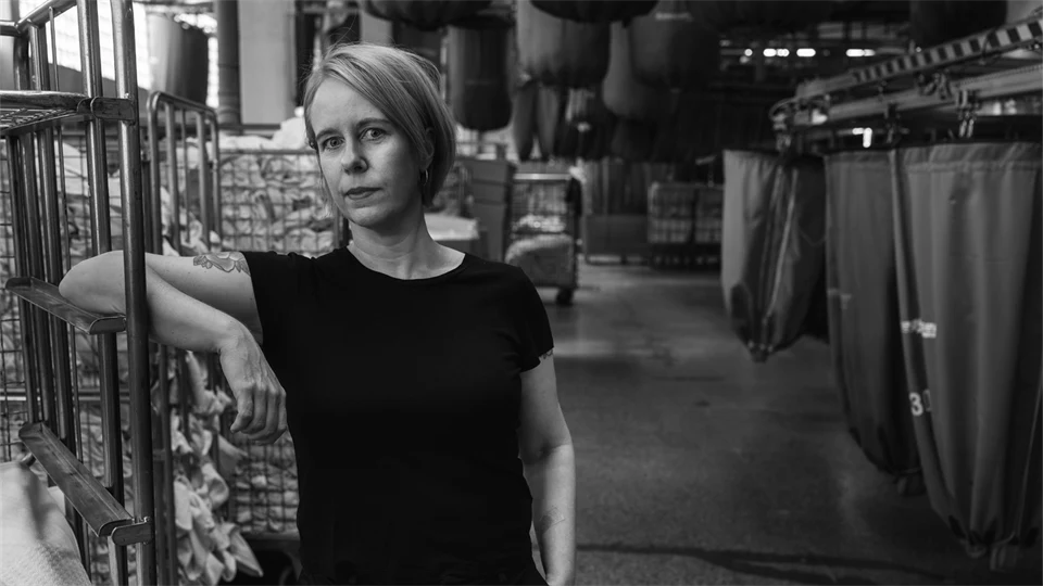 En kvinna med ljust hår, klädd i svart, poserar framför kameran, i bakgrunden syns vagnar med tvätt.