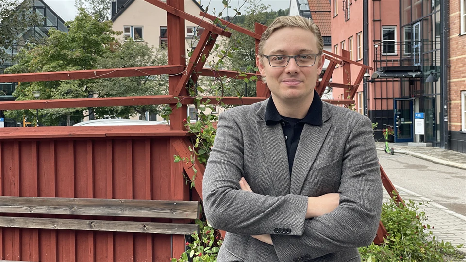 A fair-haired man with glasses and a blazer stands in front of a red fence and poses for the camera.