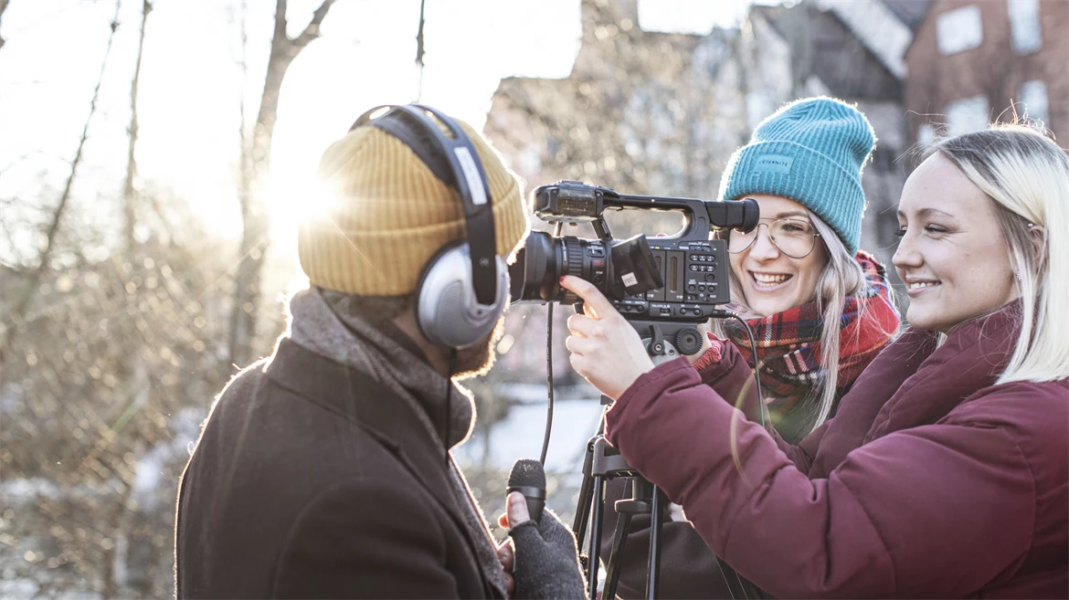 Utomhusbild med en kille med hörlurar som håller i en mikrofon. Bakom en filmkamera står två tjejer.