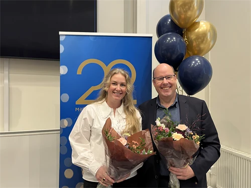 En kvinna och en man står framför ballonger. De håller i varsin blombukett och ser glada ut. 