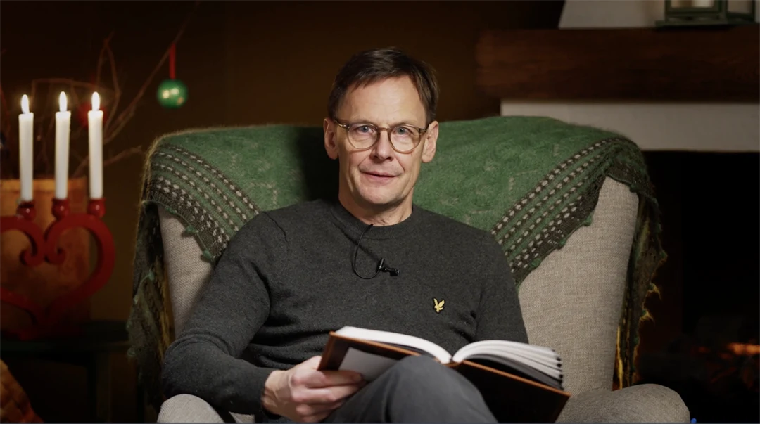 En man sitter i en fåtölj och läser en bok. I bakgrunden en öppen spis och en ljusstake.