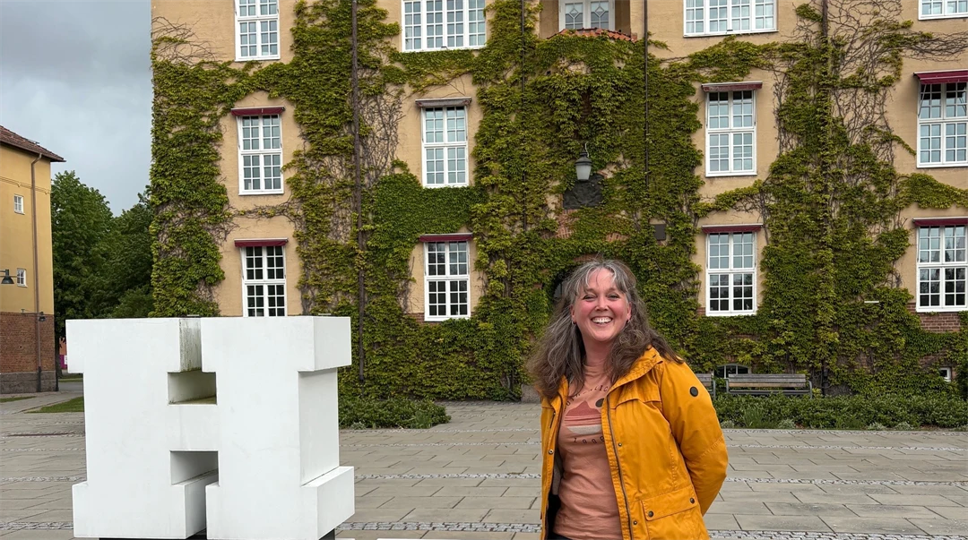 A woman in a yellow jacket is standing outside a beige building with green climbing plants. Next to her is a sculpture with the letters ‘HKR’.