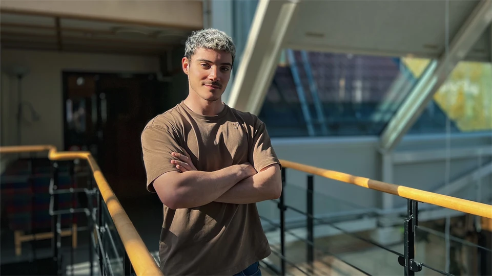  A young man in a t-shirt stands by a railing in a room with windows where the sunlight falls.