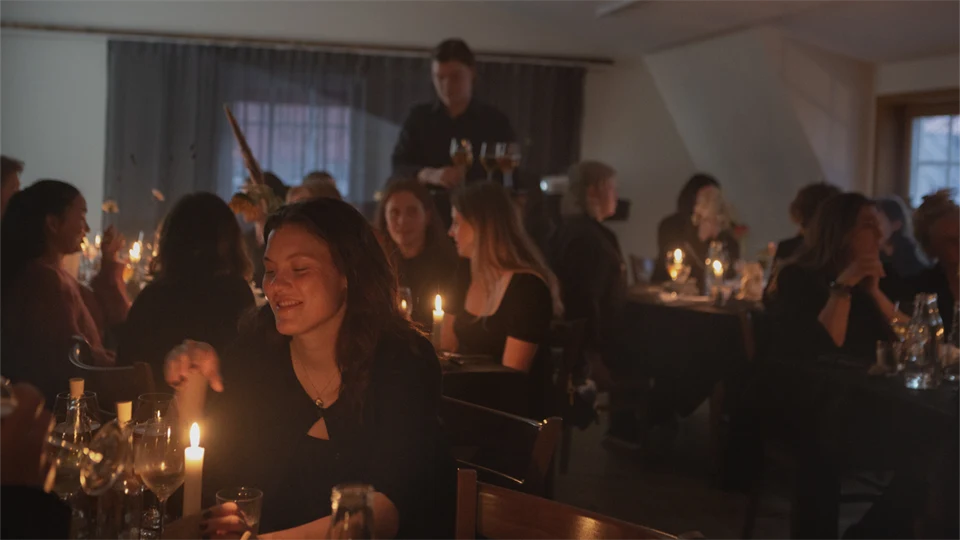 Människor sitter vid dukade bord i dunkelt rum med tända stearinljus, en person serverar drycker från en bricka.