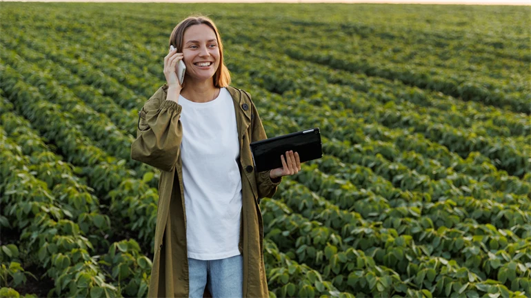 Ung kvinnlig jordbrukare står i stor odling av bönor, hon håller en telefon i ena handen och en surfplatta i den andra. Hon ser glad ut.