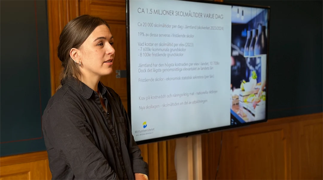 A woman with brown hair and a dark grey shirt stands by a screen with a presentation on it.