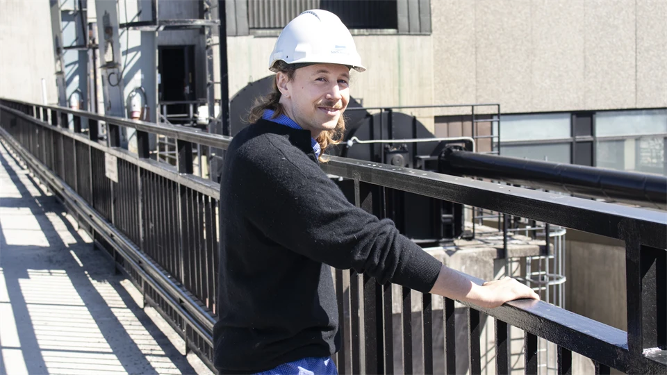  A man in a white helmet stands by a black fence in an industrial setting.