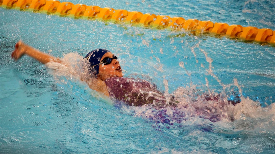 A girl wearing swimming goggles, a swimming cap and a swimsuit is swimming backstroke.