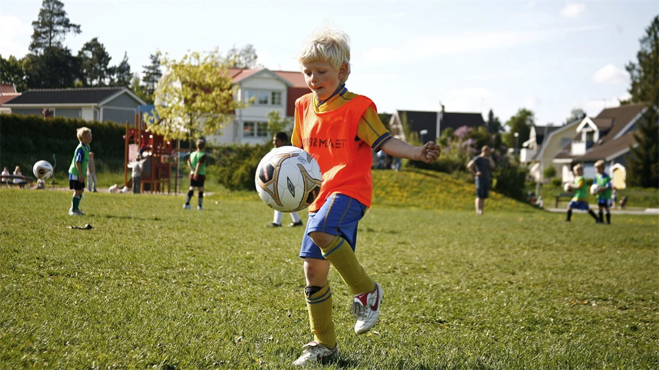 En pojke med blondt hår i fotbollskläder jonglerar med en fotboll på en gräsplan med andra barn och villor i bakgrunden. 