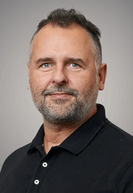 Porträttbild av en man i svart skjorta, svart hår med gråa inslag och skägg med en grå bakgrund. 