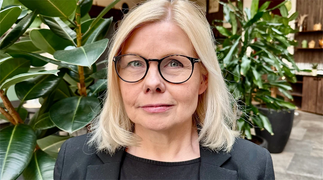 En kvinna med axellångt blont hår och glasögon, klädd i svart kavaj står framför gröna, höga växter i inomhusmiljö. 