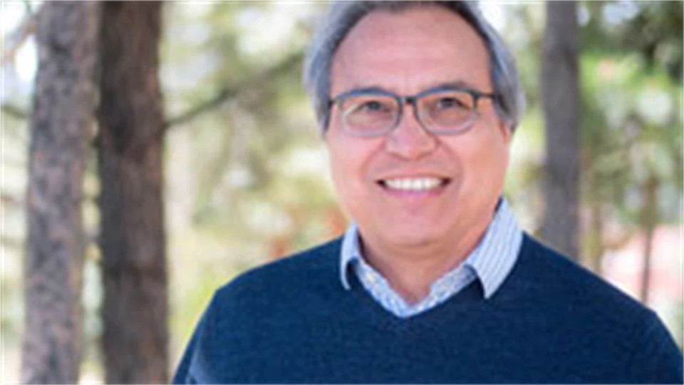 A man with glasses wearing a blue and white checkered shirt with a blue sweater over it stands outside with trees in the background