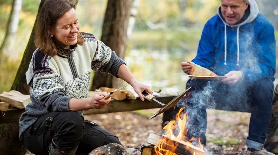 Kvinna i ulltröja och man i hoodie lagar mat över en eldstad i skogen
