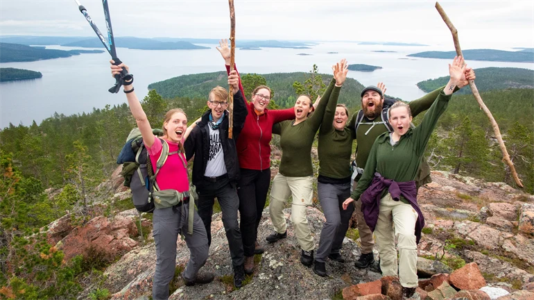 En grupp människor jublar över att ha nått toppen, i bakgrunden utsikt över skog och vatten.