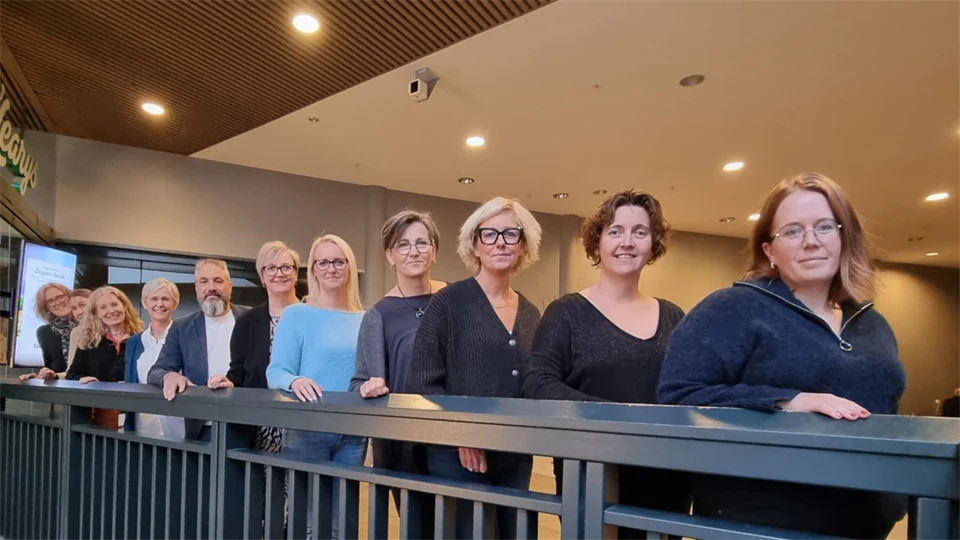 Ett tiotal kontorskläddda personer, både kvinnor och män, står uppradade längs ett räcke och tittar in i kameran. 