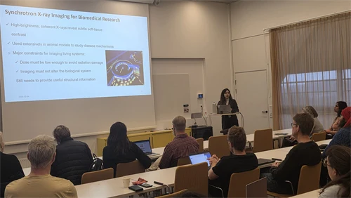 A woman stands at the front of a lecture hall with tables and chairs where the audience sits and listens. 