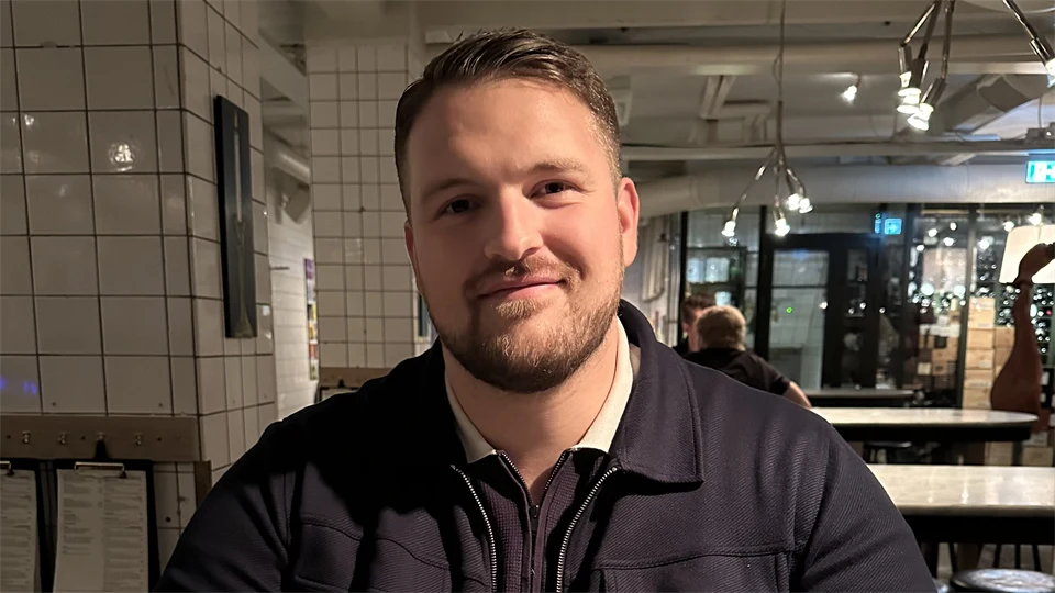 Man med mörkt kort hår, skägg och mörkblå kofta tittar in i kameran.