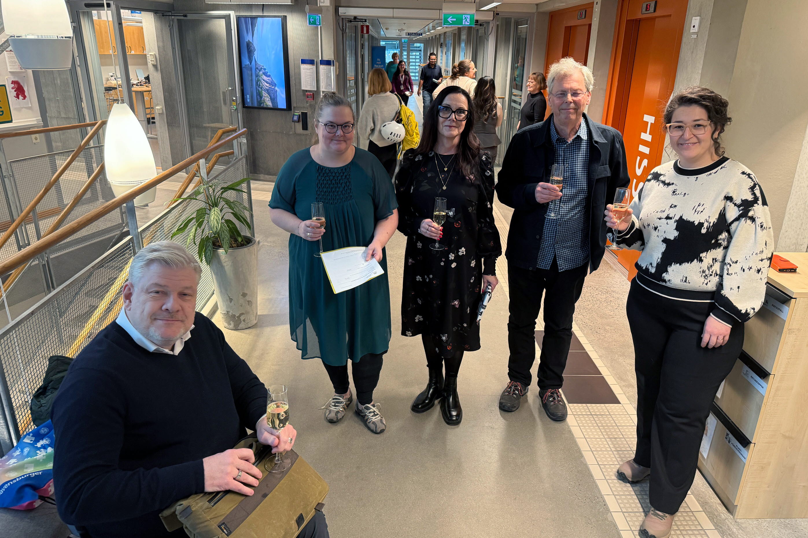 tre kvinnor och en man står tillsammans och håller i varsitt glas, en av dem siter ned