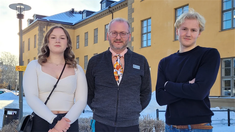 Tre personer står framför en gul byggnad med vita fönster på en snöig dag