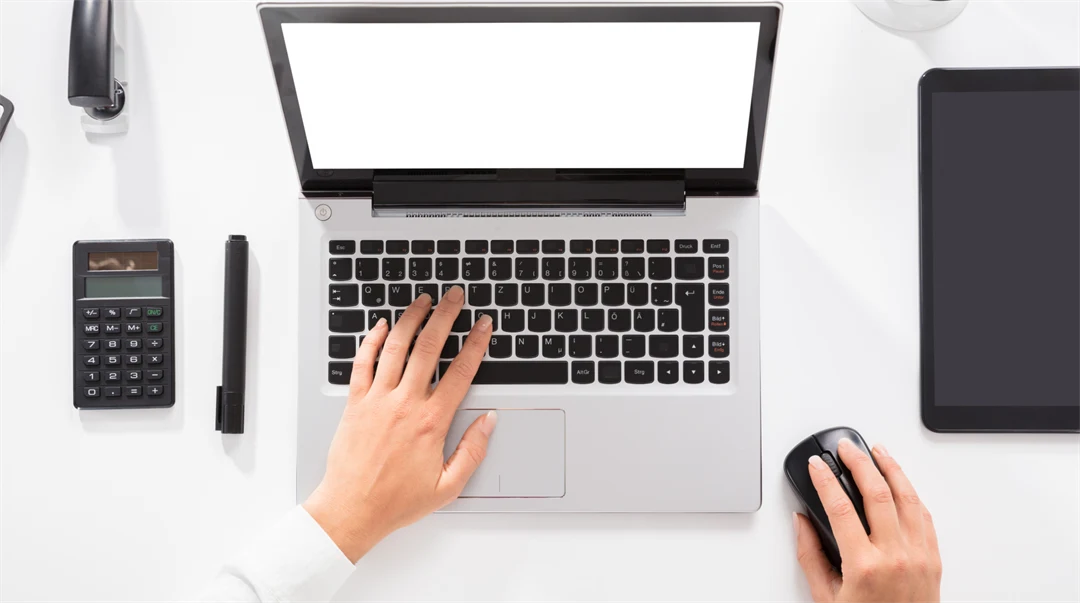 Händer knappar på en laptop, miniräknare, tuschpenna och häftmaskin ligger in till