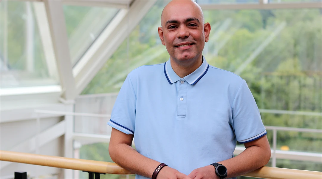 Man in blue short-sleeved shirt stands by a railing and smiles at the camera.