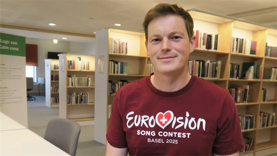 Ung kortklippt man i mörkröd t-shirt med Eurovision-tryck, sittandes vid ett bord i ett bibliotek med böcker i bakgrunden