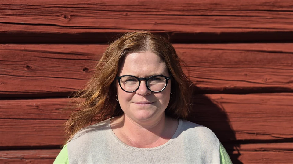 En kvinna med brunt hår och glasögon mot en röd trävägg