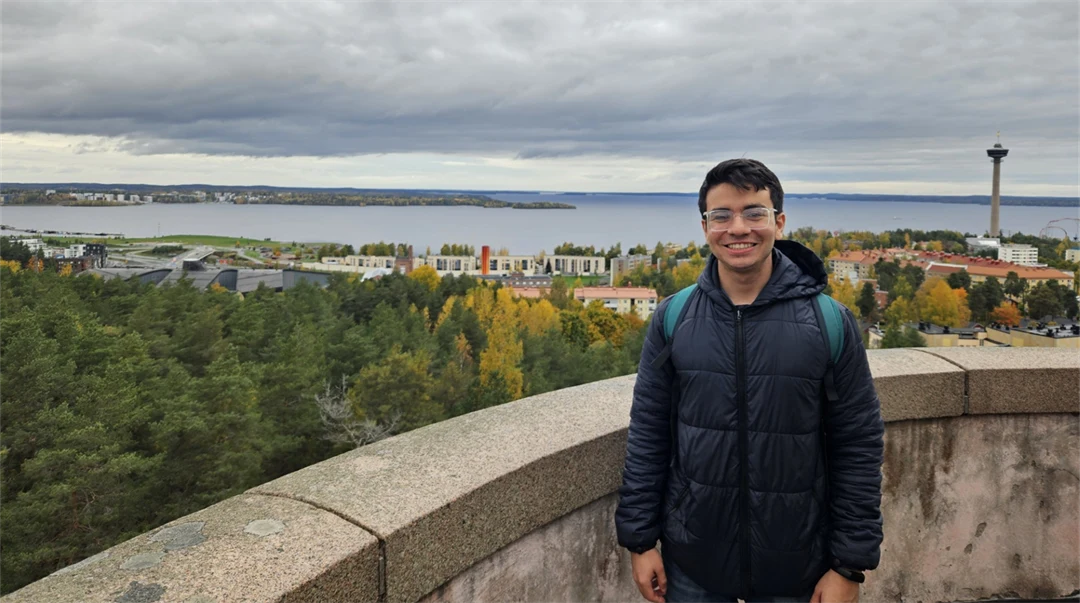 A young man wearing a dark blue jacket and carrying a rucksack is standing by a stone wall high up, looking out over trees, buildings and water. 