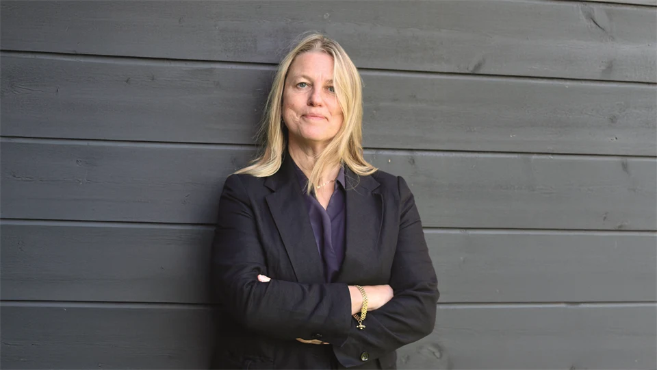 En blond kvinna står med armarna i kors framför en svart trävägg.