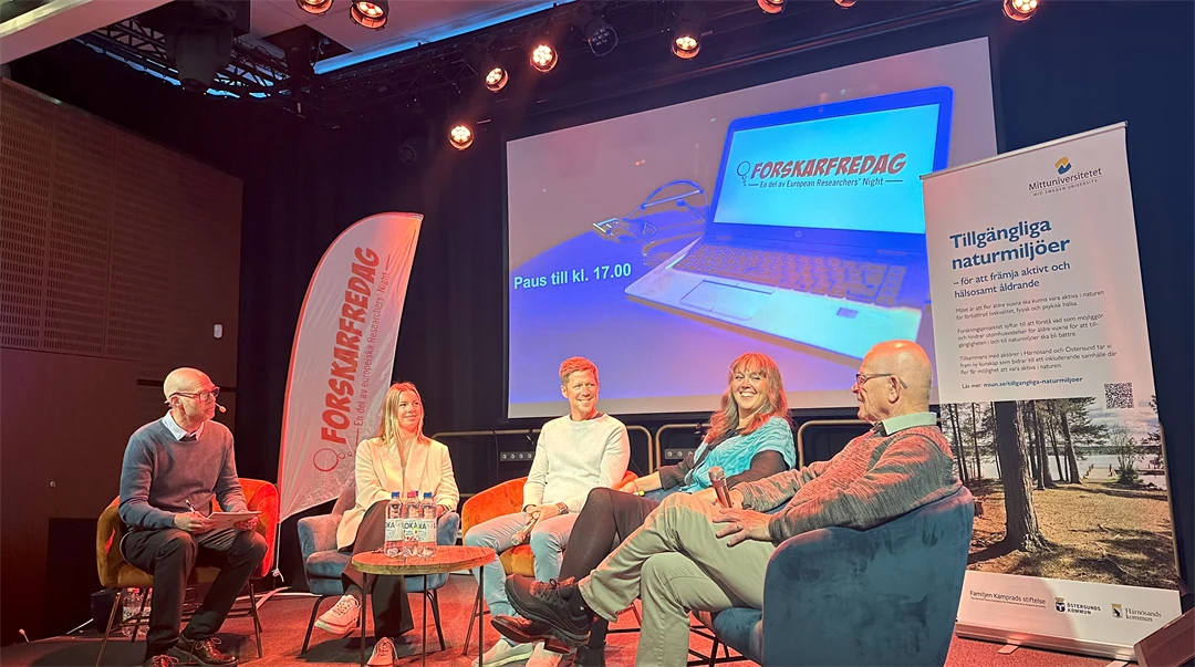 Fem personer sitter på stolar på en scen framför en skärm. 